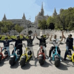 A group of eight people on motorized scooters raise their arms in celebration. They are in an urban park with historic buildings and greenery in the background.