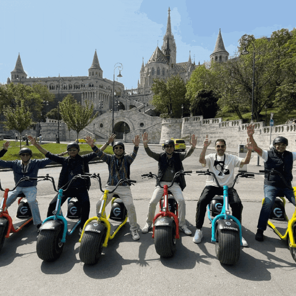 A group of eight people on motorized scooters raise their arms in celebration. They are in an urban park with historic buildings and greenery in the background.