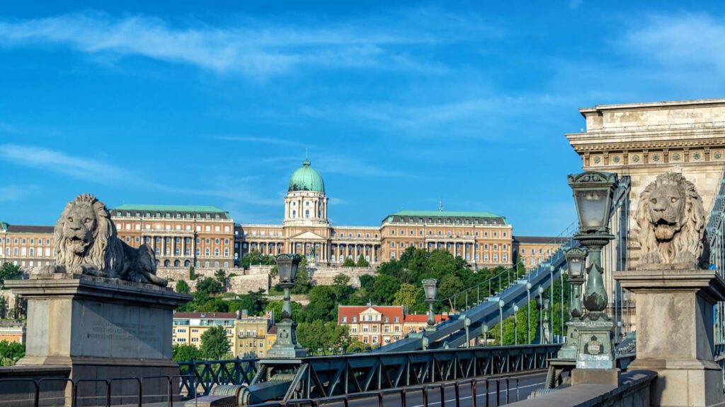 The image shows the Chain Bridge with lion statues in the foreground and Buda Castle in the background under a clear blue sky.