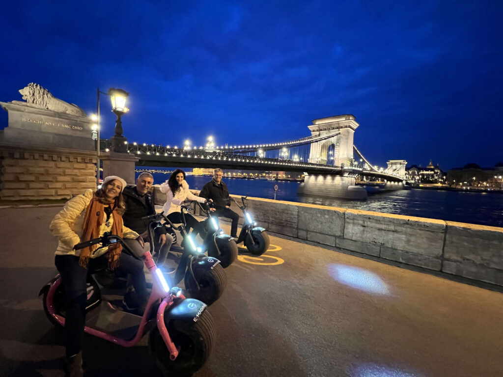 A group of people on electric scooters poses near a river at night with a lit-up bridge in the background.