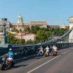 People on e-scooter tours ride over the Chain Bridge in Budapest, Hungary, with Buda Castle visible in the background on a clear day.