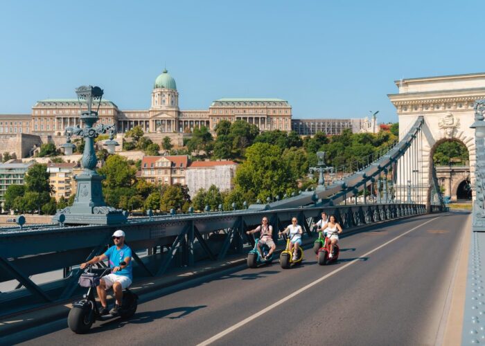 People on e-scooter tours ride over the Chain Bridge in Budapest, Hungary, with Buda Castle visible in the background on a clear day.