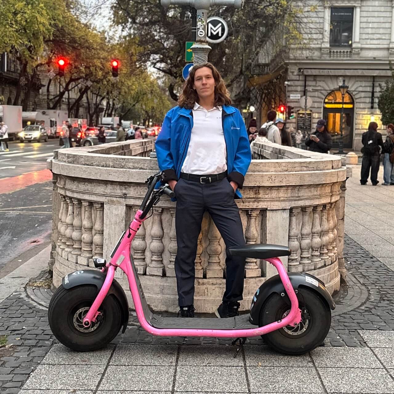 A person in a blue jacket and dark pants stands confidently with hands in pockets behind a pink electric scooter on a city street, with historic stone railings and buildings in the background.