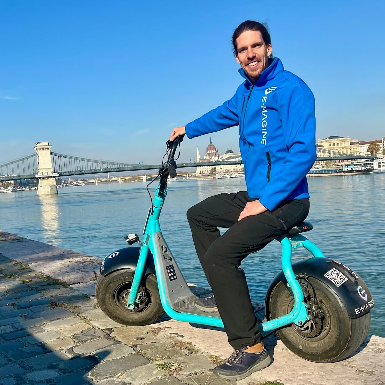 A man in a bright blue jacket sits on a blue electric scooter by a river, with a bridge and cityscape in the background under a clear blue sky.