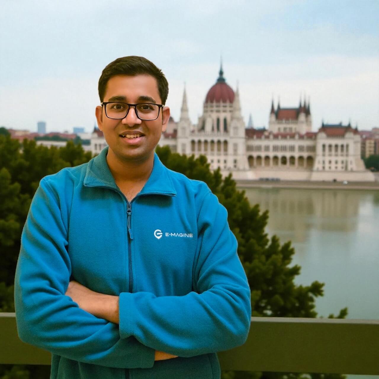 A smiling person in glasses and a blue jacket stands with arms crossed on a balcony, with a large, ornate building and river in the background. Trees are visible in the foreground.