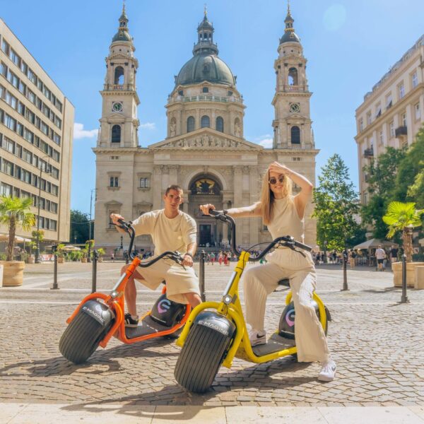 Két ember ül elektromos robogón egy nagy, ikertornyos és kupolás történelmi épület előtt, megörökítve Budapest napfényes, macskaköves plázájának eszenciáját.
