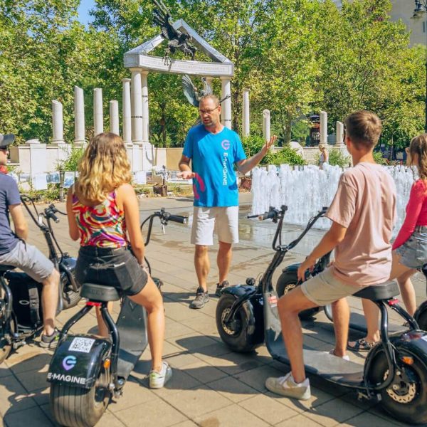 A tour guide wearing a blue shirt speaks to four people seated on electric scooters in front of a fountain and pillars, in a park setting with trees in the background.