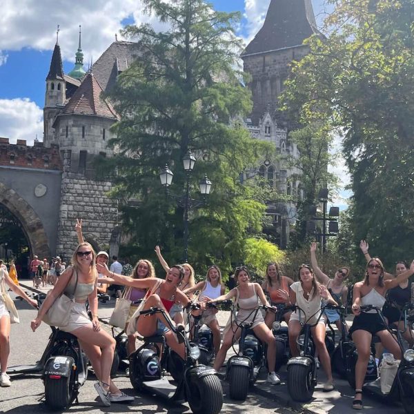 A group of people on electric scooters posing for a photo in front of a historic castle gate under a clear sky.