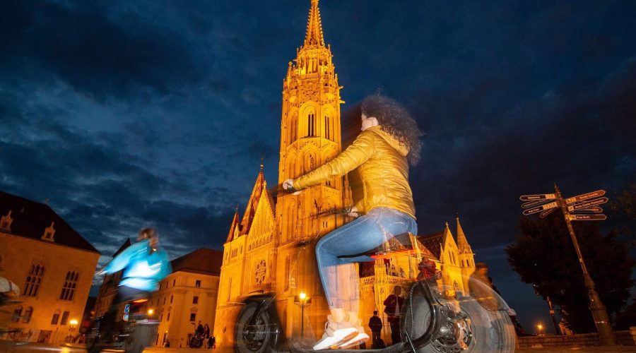People riding bicycles near an illuminated Gothic-style cathedral at dusk.
