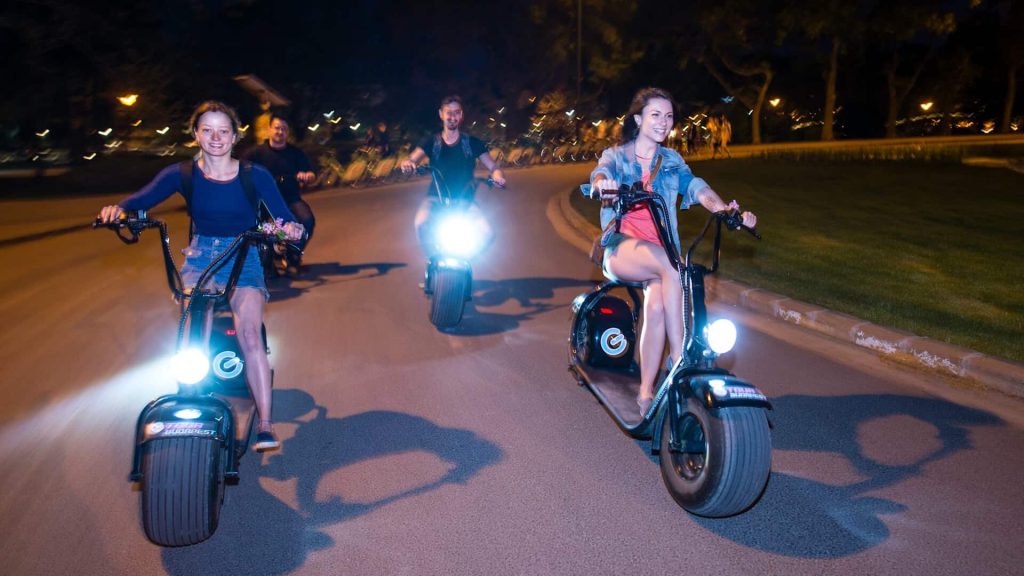 Four people ride electric scooters on a road at night, smiling and appearing to enjoy themselves. The headlights of the scooters are on, illuminating the path ahead.