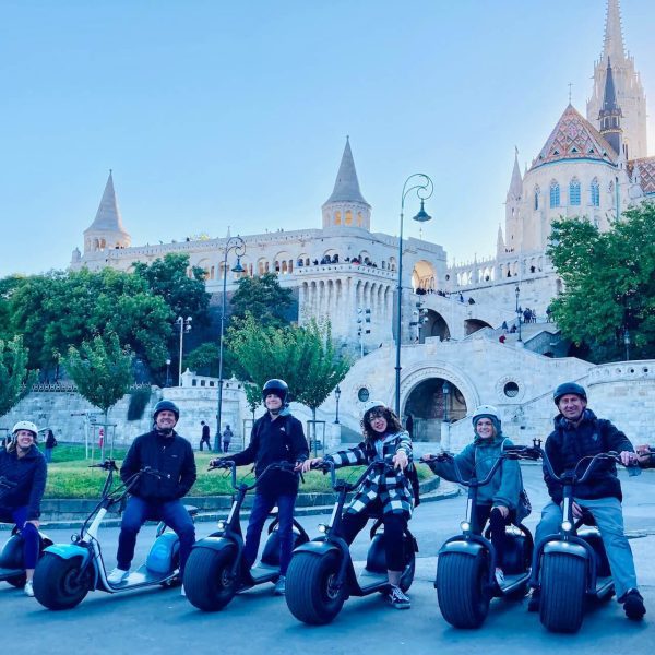 Hétfős csoport elektromos robogókon pózol egy történelmi épület és kastélyszerű építmény előtt. Nappal van.