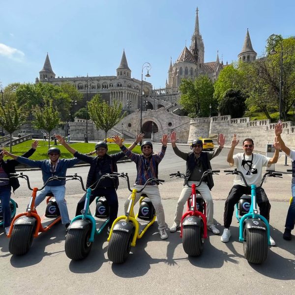 A group of eight people on electric scooters raise their arms in front of a historic building with multiple towers and a staircase, showcasing the joy of discovering where and how to rent an electric scooter in Budapest.