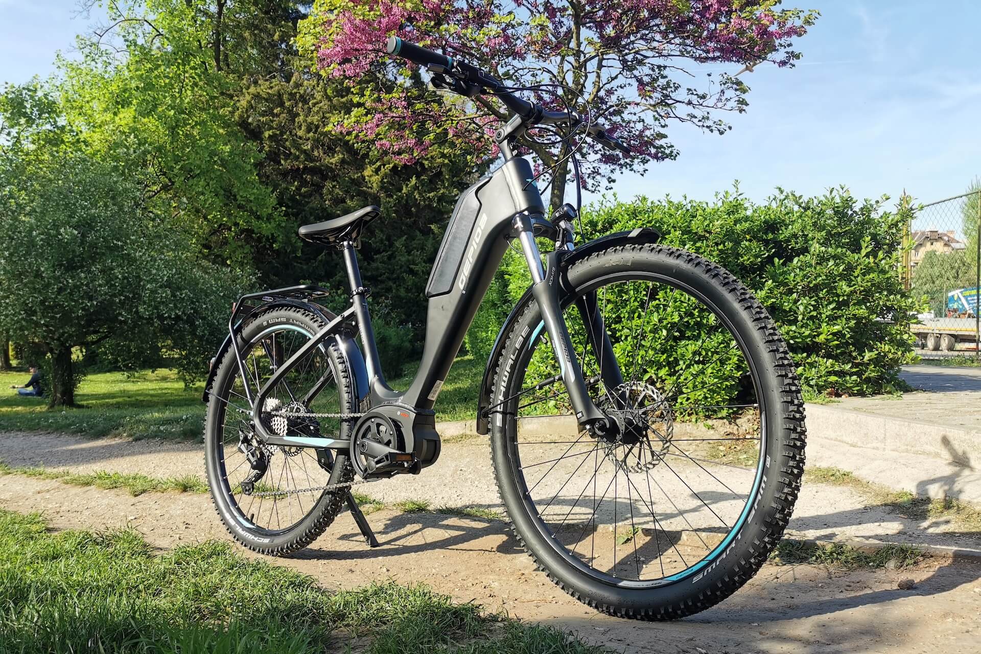 Egy fekete elektromos kerékpár parkol egy ösvényen egy parkban, különböző zöld fákkal és bokrokkal a háttérben.