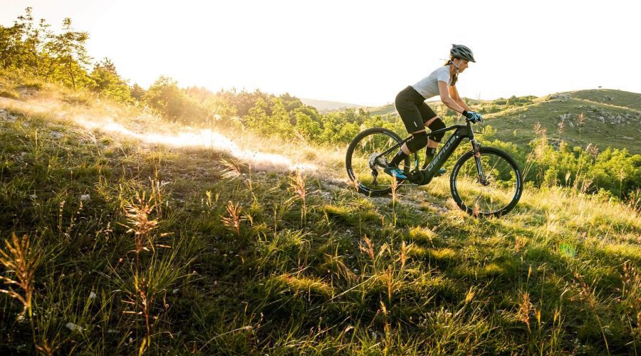Egy sisakot viselő személy mountain bike-on megy le egy füves dombról napkeltekor vagy napnyugtakor.