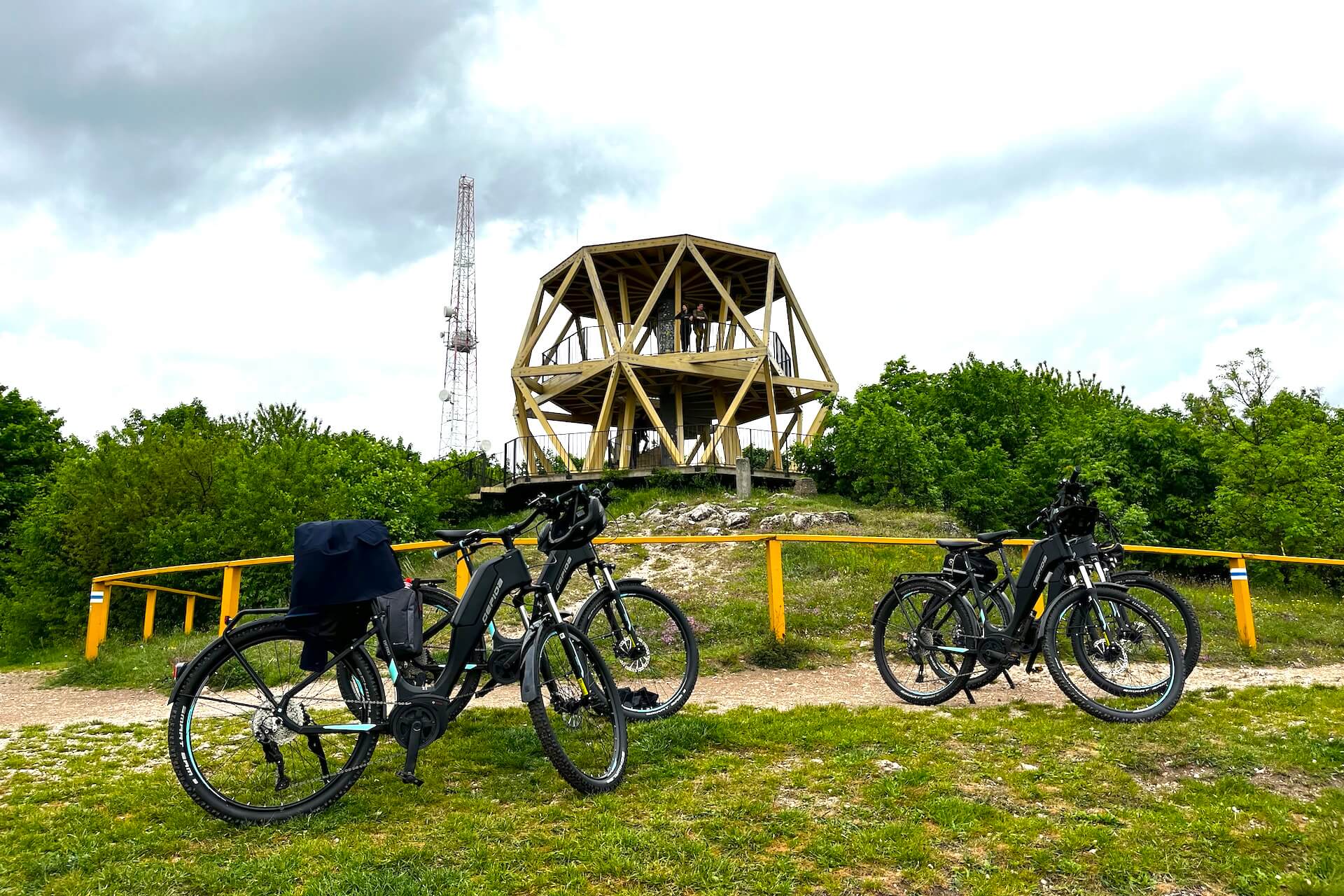 Három kerékpár parkol egy fából készült kilátó előtti füves területen. A háttérben egy kommunikációs torony áll, zöldövezetben, felhős ég alatt.