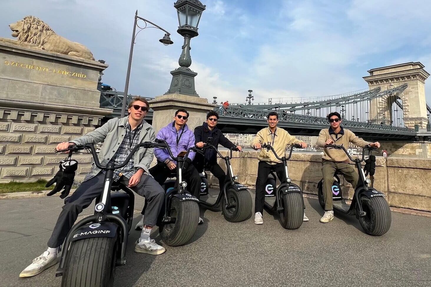 Five people pose on electric scooters in front of the iconic Chain Bridge in Budapest on a sunny day—truly the best way to explore this vibrant city.