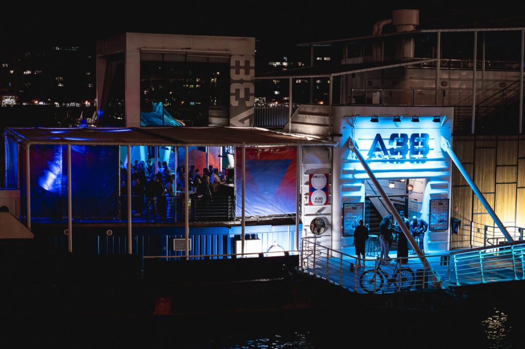 A modern, brightly lit riverside bar or club in Budapest at night, illuminated with blue and purple lights. People gather on the outdoor deck while bicycles are parked near the entrance—a must-see spot for any travel guide of things to do.