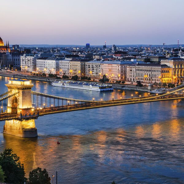 A twilight view of Budapest’s Chain Bridge spanning the Danube River, with city lights reflecting on the water and the Hungarian Parliament building illuminated in the background—a must-see highlight in any Budapest guide.