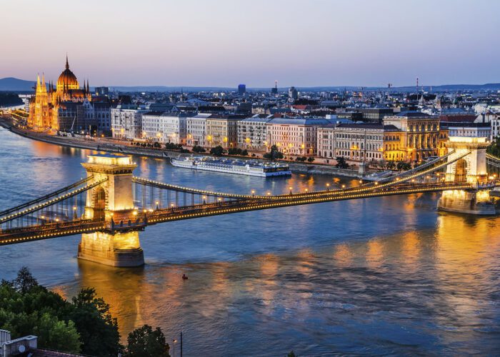 A twilight view of Budapest’s Chain Bridge spanning the Danube River, with city lights reflecting on the water and the Hungarian Parliament building illuminated in the background—a must-see highlight in any Budapest guide.