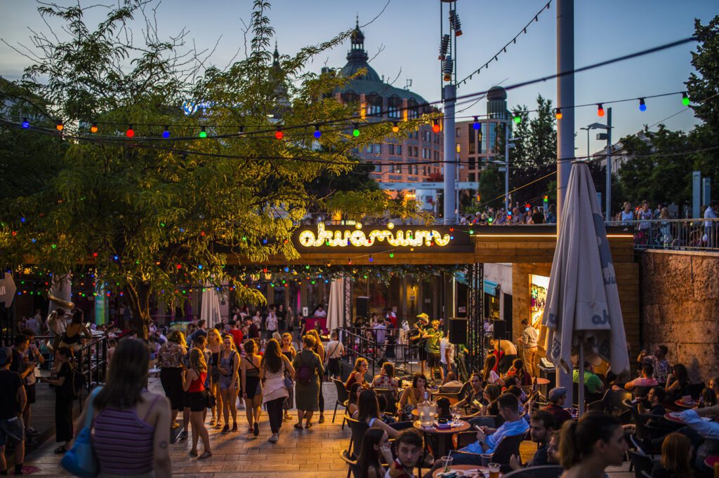 A lively outdoor plaza at dusk, crowded with people socializing and walking—an ideal spot for those seeking things to do in Budapest. Colorful string lights shine overhead, a glowing sign reads obuvarium, with trees and historic buildings in the background.
