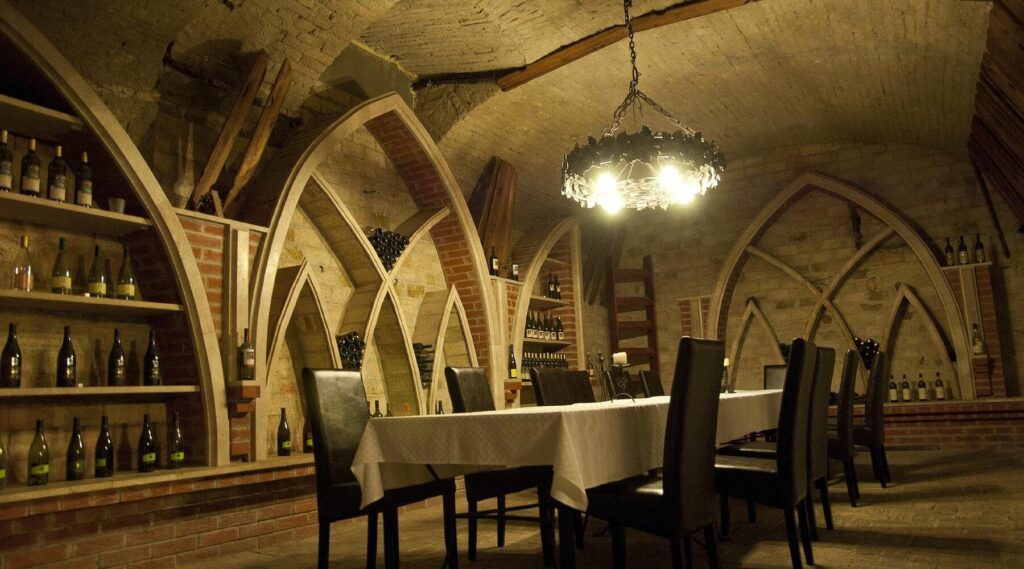 A dimly lit wine cellar with arched brick ceilings, shelves of wine bottles, and a long dining table beneath an ornate chandelier—an atmospheric spot that tops many Budapest at night lists for unique things to do in Budapest.