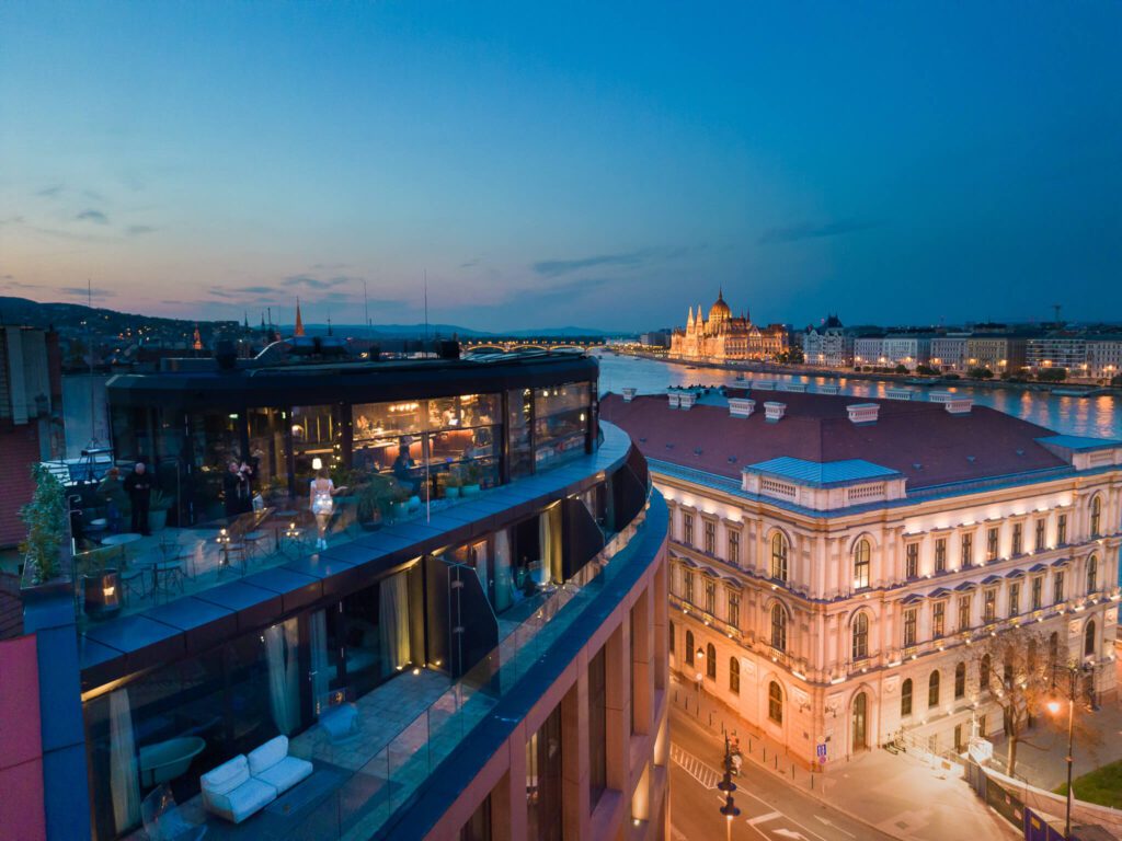 A rooftop bar with glass walls overlooks Budapest at night, offering stunning views of the illuminated Hungarian Parliament and Chain Bridge across the Danube River—an unforgettable highlight in any Budapest guide.