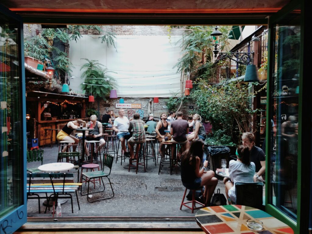 People sit at tables in a cozy outdoor courtyard cafe with plants, string lights, and eclectic decor—an ideal spot for things to do in Budapest at night, where groups eat, talk, and unwind in the lively, casual atmosphere.