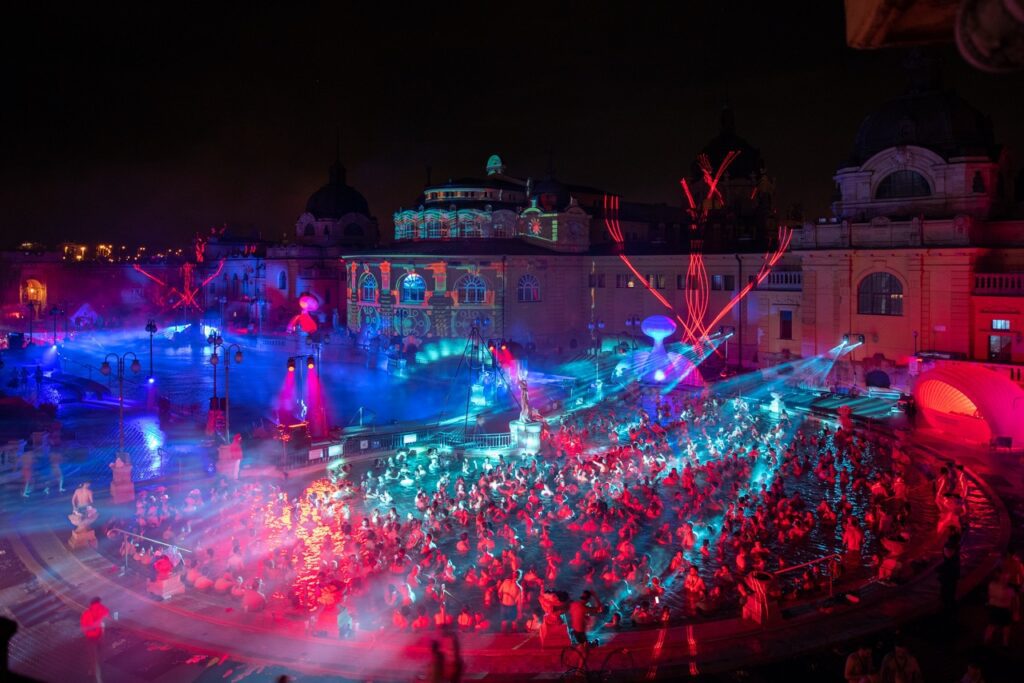 A large crowd gathers in a historic outdoor pool at night, illuminated by vibrant red and blue lights and laser beams, during a festive event—one of the top things to do in Budapest. The ornate building behind glows with colorful projections.